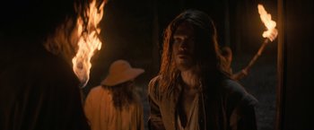 Movie still from “Fear Street: Part Three - 1666” (2021), directed by Leigh Janiak – A man with long hair standing in front of a fire; Medium shot, Over the shoulder angle
