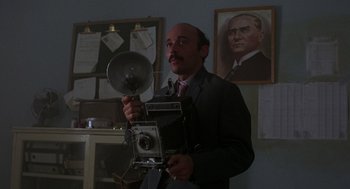 Movie still from “Midnight Express” (1978), directed by Alan Parker – A man in a suit holding an old fashioned camera; Close Up shot, Low angle