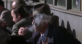 Movie still from “Midnight Express” (1978), directed by Alan Parker – An older man in a suit and hat smoking a cigarette; Close Up shot, Over the shoulder angle
