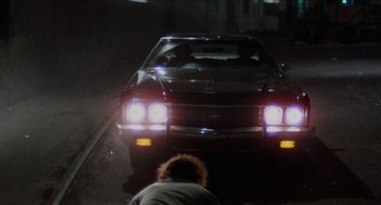 Movie still from “Mikey and Nicky” (1976), directed by Elaine May – A man kneeling down in front of a parked car at night; Wide shot, High angle