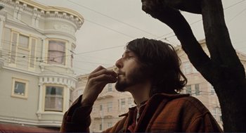 Movie still from “Milk” (2008), directed by Gus Van Sant – A man eating a donut while standing next to a building; Medium shot, Low angle