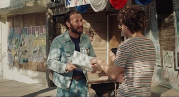 Movie still from “Milk” (2008), directed by Gus Van Sant – A man in a denim jacket is handing something to another man in a striped shirt; Medium shot, Over the shoulder angle