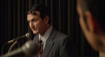 Movie still from “Milk” (2008), directed by Gus Van Sant – A man wearing a suit and tie talking into a microphone; Close Up shot, Over the shoulder angle