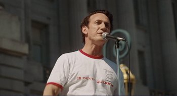 Movie still from “Milk” (2008), directed by Gus Van Sant – A man in a white t - shirt is holding a microphone; Medium shot, Low angle