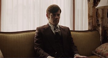 Movie still from “Milk” (2008), directed by Gus Van Sant – A man sitting on a couch wearing a suit and tie; Medium shot, Over the shoulder angle