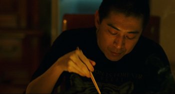 Movie still from “Millennium Mambo” (2001), directed by Hsiao-Hsien Hou – A man holding chopsticks while sitting in a chair; Close Up shot, Over the shoulder angle