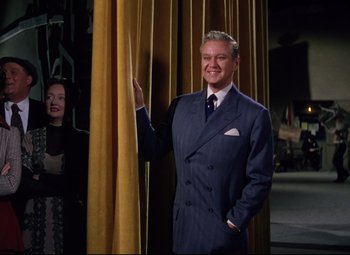 Movie still from “Million Dollar Mermaid” (1952), directed by Mervyn LeRoy – A man wearing a suit and tie standing next to a curtain; Medium shot, Low angle
