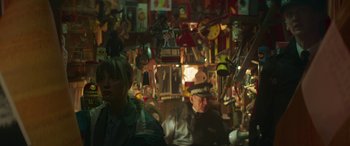Movie still from “Mindhorn” (2016), directed by Sean Foley – A man and a woman sitting at a table with a lot of toys hanging on the wall; Medium shot, Over the shoulder angle