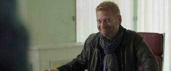 Movie still from “Mindhorn” (2016), directed by Sean Foley – A man wearing a jacket and a scarf smiling for the camera; Close Up shot, Over the shoulder angle