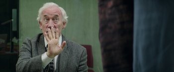 Movie still from “Mindhorn” (2016), directed by Sean Foley – An older man in a gray suit and white hair is holding his hand up to his face; Close Up shot, Over the shoulder angle