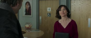Movie still from “Mindhorn” (2016), directed by Sean Foley – A woman sitting at a table in front of a restroom door; Medium shot, Over the shoulder angle