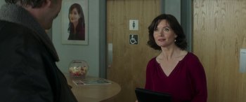 Movie still from “Mindhorn” (2016), directed by Sean Foley – A woman sitting at a table in front of a restroom sign; Medium shot, Over the shoulder angle
