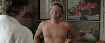 Movie still from “Mindhorn” (2016), directed by Sean Foley – A man with no shirt is smiling for the camera; Medium shot, Over the shoulder angle