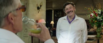 Movie still from “Mindhorn” (2016), directed by Sean Foley – A man holding a wine glass and smiling at another man; Medium shot, Over the shoulder angle