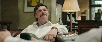 Movie still from “Mindhorn” (2016), directed by Sean Foley – A man sitting on top of a brown leather couch; Medium shot, Low angle