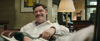 Movie still from “Mindhorn” (2016), directed by Sean Foley – A man sitting in a chair smiling at the camera; Medium shot, Over the shoulder angle