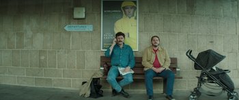 Movie still from “Mindhorn” (2016), directed by Sean Foley – Two men sitting on a wooden bench in front of a poster; Medium shot, Low angle