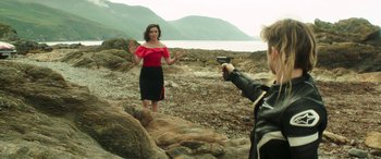 Movie still from “Mindhorn” (2016), directed by Sean Foley – A woman standing on a rocky shore while a man takes a picture; Medium shot, Over the shoulder angle