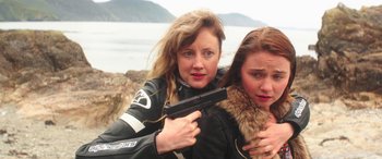 Movie still from “Mindhorn” (2016), directed by Sean Foley – Two women are posing for a picture while holding a gun; Close Up shot, Low angle
