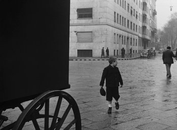 Movie still from “Miracle in Milan” (1951), directed by Vittorio De Sica – A boy walking down the street in front of a building; Wide shot, Low angle