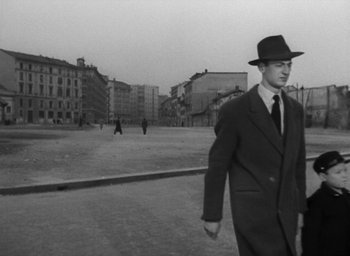 Movie still from “Miracle in Milan” (1951), directed by Vittorio De Sica – A black and white photo of a man in a suit and hat; Wide shot, Low angle