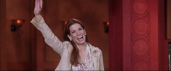 Movie still from “Miss Congeniality 2: Armed & Fabulous” (2005), directed by John Pasquin – A woman in a pink dress and a jacket is holding her arm up; Medium shot, High angle