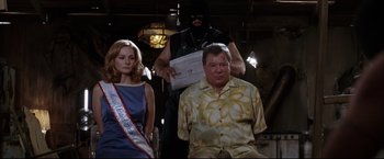Movie still from “Miss Congeniality 2: Armed & Fabulous” (2005), directed by John Pasquin – A man wearing a mask is sitting next to a woman in a blue dress; Medium shot, Overhead angle