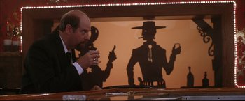 Movie still from “Miss Congeniality 2: Armed & Fabulous” (2005), directed by John Pasquin – A man sitting at a table in front of a shadow of a cowboy; Medium shot, Low angle