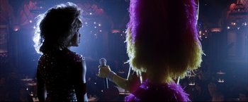 Movie still from “Miss Congeniality 2: Armed & Fabulous” (2005), directed by John Pasquin – A person holding a microphone and a person in a purple costume; Close Up shot, Over the shoulder angle