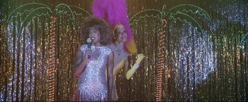 Movie still from “Miss Congeniality 2: Armed & Fabulous” (2005), directed by John Pasquin – Two women dressed up in costumes singing into microphones in front of lights; Medium shot, Over the shoulder angle