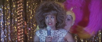 Movie still from “Miss Congeniality 2: Armed & Fabulous” (2005), directed by John Pasquin – A woman in a silver sequined dress holding a microphone; Close Up shot, Over the shoulder angle