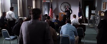 Movie still from “Miss Congeniality 2: Armed & Fabulous” (2005), directed by John Pasquin – A man standing at a podium in front of a group of people; Wide shot, Over the shoulder angle