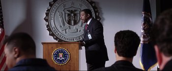 Movie still from “Miss Congeniality 2: Armed & Fabulous” (2005), directed by John Pasquin – A man standing at a podium in front of an fbi logo; Medium shot, Low angle