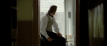 Movie still from “Mister Lonely” (2007), directed by Harmony Korine – A man sitting on a chair in front of a window; Medium shot, Over the shoulder angle