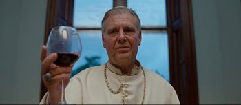 Movie still from “Mister Lonely” (2007), directed by Harmony Korine – A man in a priest's outfit holding a glass of wine in front of a window; Close Up shot, Low angle