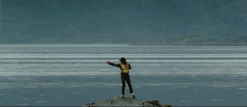 Movie still from “Mister Lonely” (2007), directed by Harmony Korine – A man standing on top of a rock in front of a body of water; Extreme Wide shot, High angle