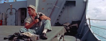 Movie still from “Mister Roberts” (1955), directed by John Ford – A man sitting on the ground holding a camera; Medium shot, High angle