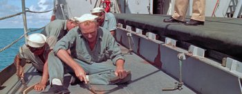 Movie still from “Mister Roberts” (1955), directed by John Ford – A man in a green shirt and a white hat is working on a boat; Medium shot, High angle