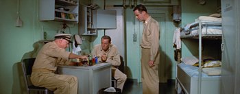 Movie still from “Mister Roberts” (1955), directed by John Ford – A man sitting at a table in a room with two other men standing next to him; Medium shot, High angle