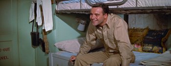 Movie still from “Mister Roberts” (1955), directed by John Ford – A man sitting on top of a bed in a room; Medium shot, High angle