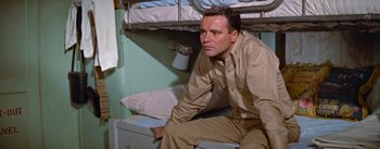 Movie still from “Mister Roberts” (1955), directed by John Ford – A man sitting on top of a bed in a room; Medium shot, High angle