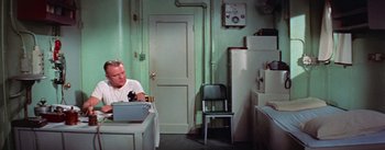 Movie still from “Mister Roberts” (1955), directed by John Ford – A man sitting at a table in front of an oven; Wide shot, High angle