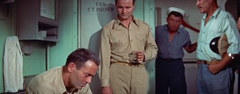 Movie still from “Mister Roberts” (1955), directed by John Ford – A man standing next to another man holding a drink; Medium shot, High angle