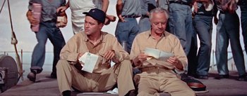 Movie still from “Mister Roberts” (1955), directed by John Ford – A couple of men sitting next to each other holding envelopes; Medium shot, Low angle
