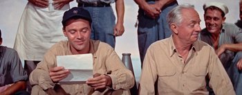 Movie still from “Mister Roberts” (1955), directed by John Ford – A man sitting next to another man while holding a piece of paper; Medium shot, Over the shoulder angle