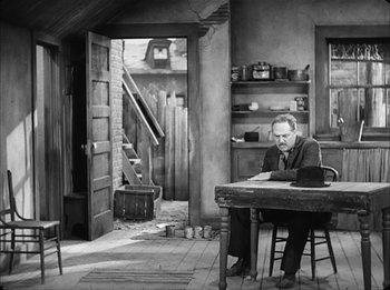 Movie still from “Modern Times” (1936), directed by Charles Chaplin – A black and white photo of a man sitting at a table; Wide shot, High angle