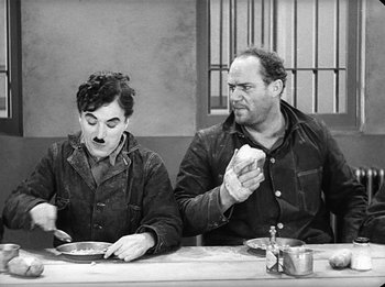 Movie still from “Modern Times” (1936), directed by Charles Chaplin – A couple of men sitting at a table eating food; Medium shot, High angle