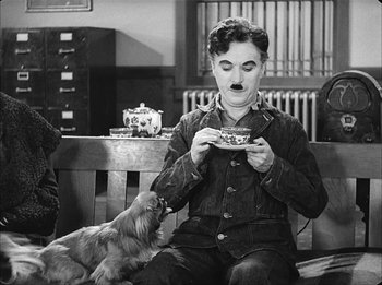 Movie still from “Modern Times” (1936), directed by Charles Chaplin – A man sitting on top of a wooden bench with a dog; Medium shot, High angle