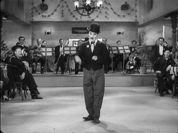 Movie still from “Modern Times” (1936), directed by Charles Chaplin – A black and white photo of a man standing in front of a orchestra; Wide shot, Low angle