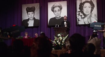 Movie still from “Mommie Dearest” (1981), directed by Frank Perry – A man standing at a podium in front of a group of people; Medium shot, Low angle